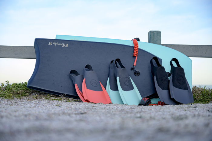 RYD Everyday swim flippers in Grey, Aqua and Coral leaning against a softboard