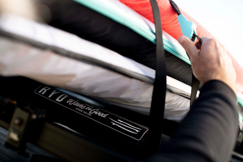Close-up of a hand tightening a black tie-down strap securing red and turquoise surfboards to a roof rack, RYD 'Waves be Good' branded pad in the foreground