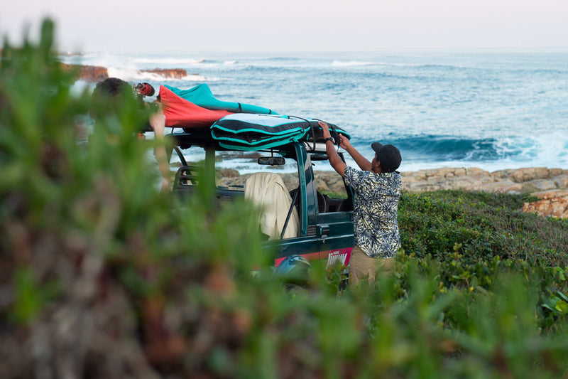 A man lifting a red and turquoise RYD board cover onto a 4x4's roof rack beside the ocean, already loaded with a padded board bag