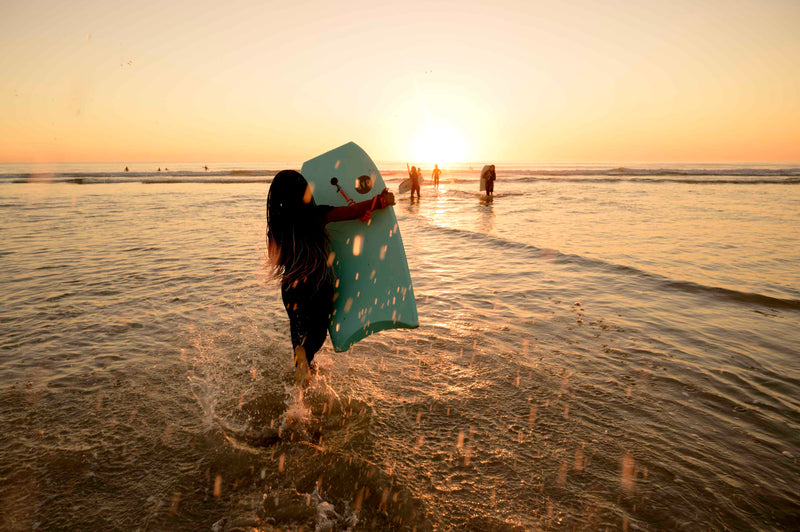 A girl running into the shorebreak at sunset carrying a mint green RYD bodyboard, other surfers silhouetted in the golden light behind her