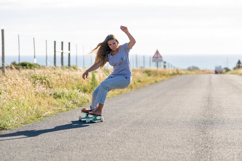 A girl carving on an RYD Everyday cruiser skateboard with turquoise wheels along a quiet coastal road, hair blowing in the wind