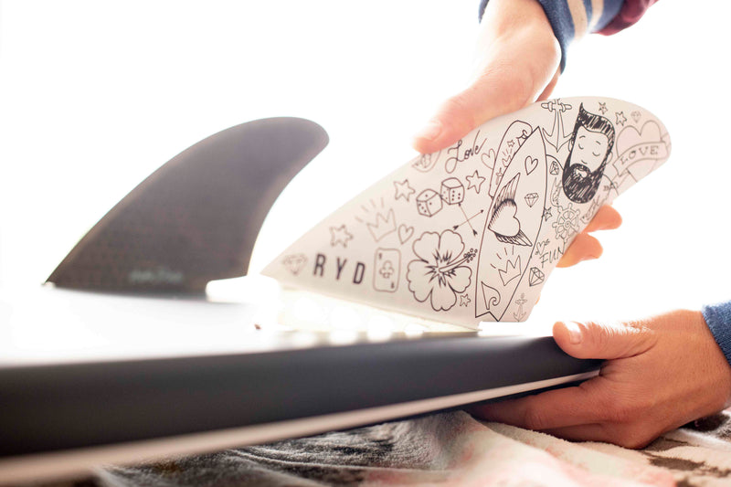 A hand fitting a RYD surfboard fin printed with a whimsical tattoo-style illustration — hibiscus, dice and a bearded surfer — into a fin box