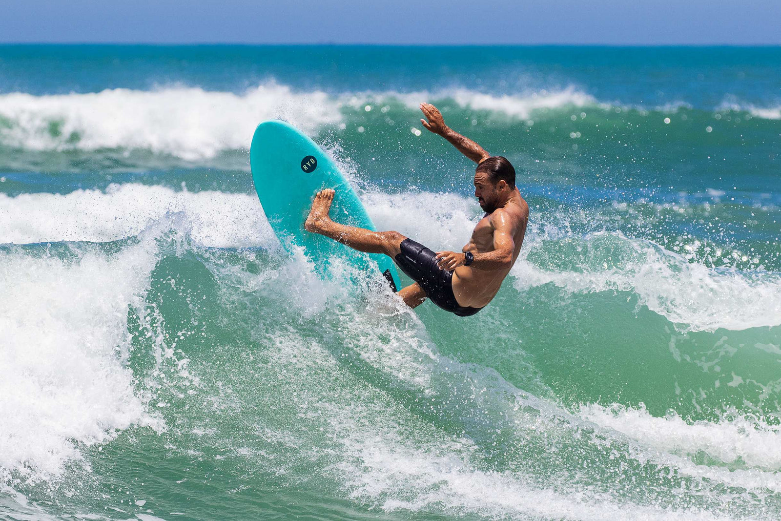 A surfer in dark boardshorts pulling a full-rotation air off the lip of a green wave on a turquoise RYD softboard, spraying whitewash, ocean and horizon behind