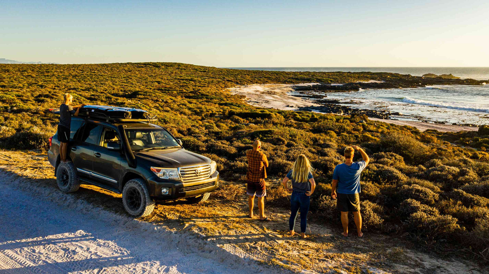 A group of friends and a lifted 4x4 with a rooftop tent parked on a coastal dirt track, looking out over fynbos scrubland toward a rocky beach at golden hour