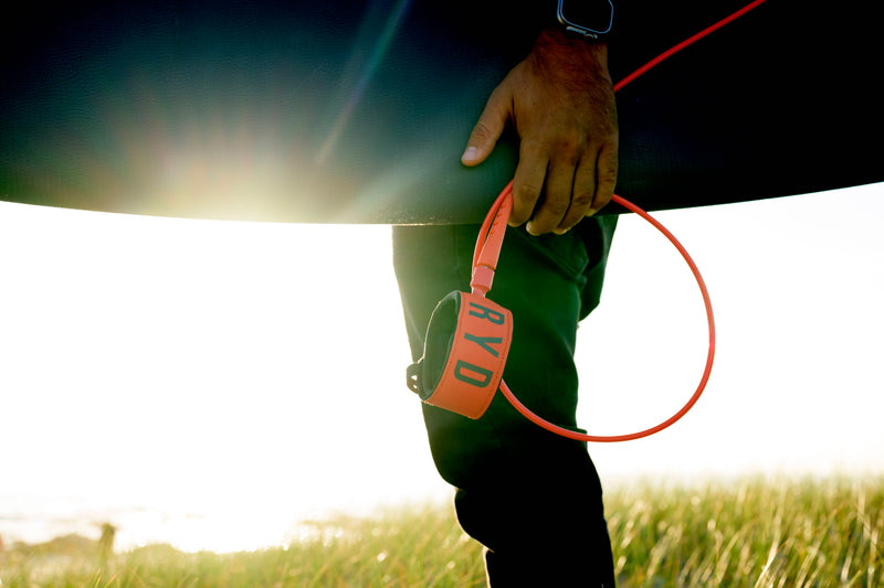 A surfer's hand holding a coiled orange RYD ankle leash cuff, wetsuit-clad leg and surfboard rail visible against a grassy dune