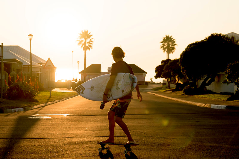 A young surfer skateboarding down a sunset street while carrying his surfboard under one arm, palm trees silhouetted against the sky