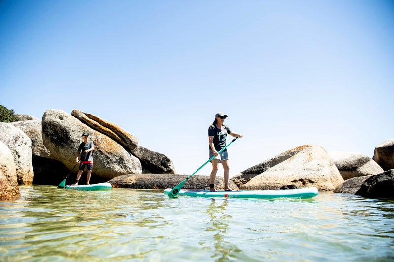 Two paddlers standing on mint green RYD Land to Sea stand-up paddleboards, weaving through large granite boulders in clear shallow water