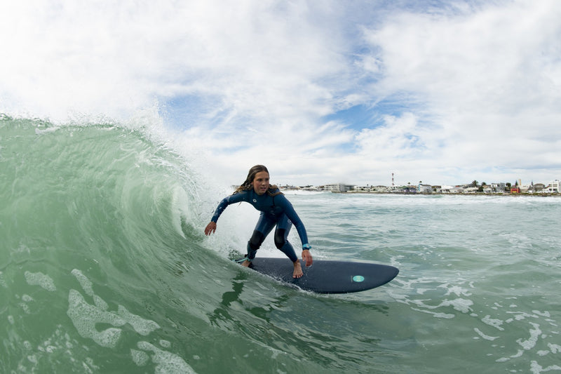 A young surfer crouched low riding inside a curling green wave on a dark RYD softboard, coastal town visible in the distance