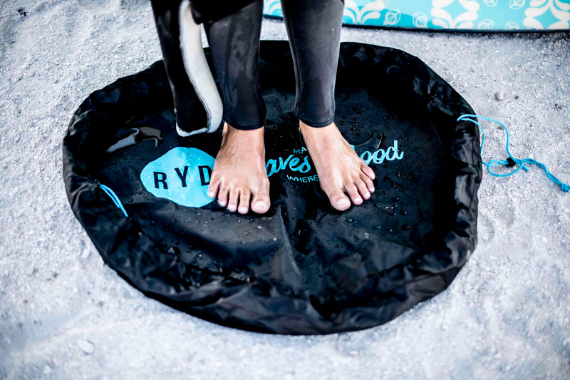 A surfer's bare feet standing on a black RYD Change Mat printed with 'May the Waves be Good Where You Are', wetsuit and surfboard beside them