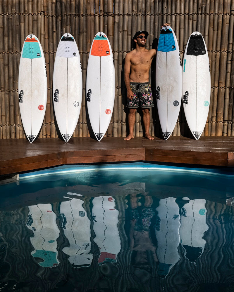 Six RYD surfboards with colour-matched traction pads standing against a bamboo wall, a surfer smiling between two of them beside a pool