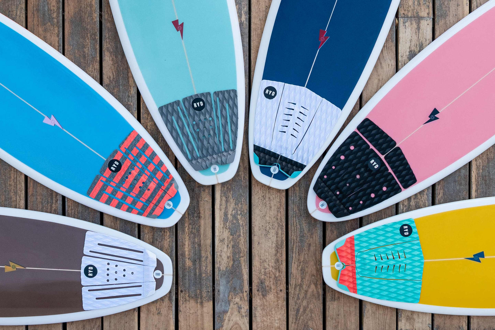 Overhead flat-lay of six surfboards fanned out, each showing a different coloured traction pad design at the tail