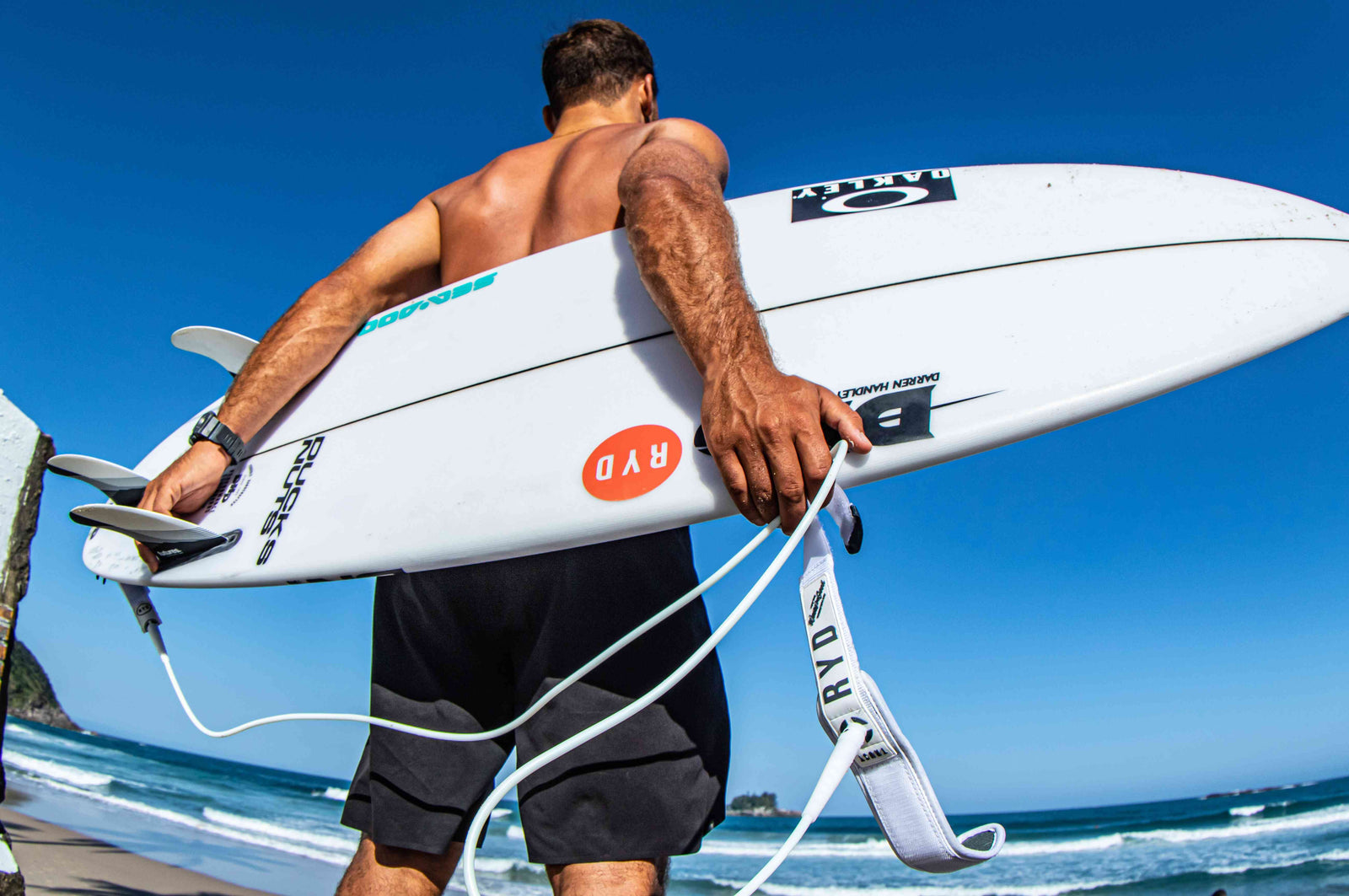 A man carrying a white surfboard under his arm on a beach, gripping its coiled leash cuff