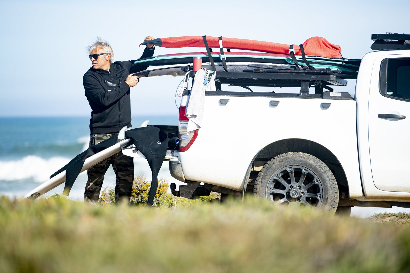 A man loading surfboards onto the roof rack of a white bakkie parked beside the ocean