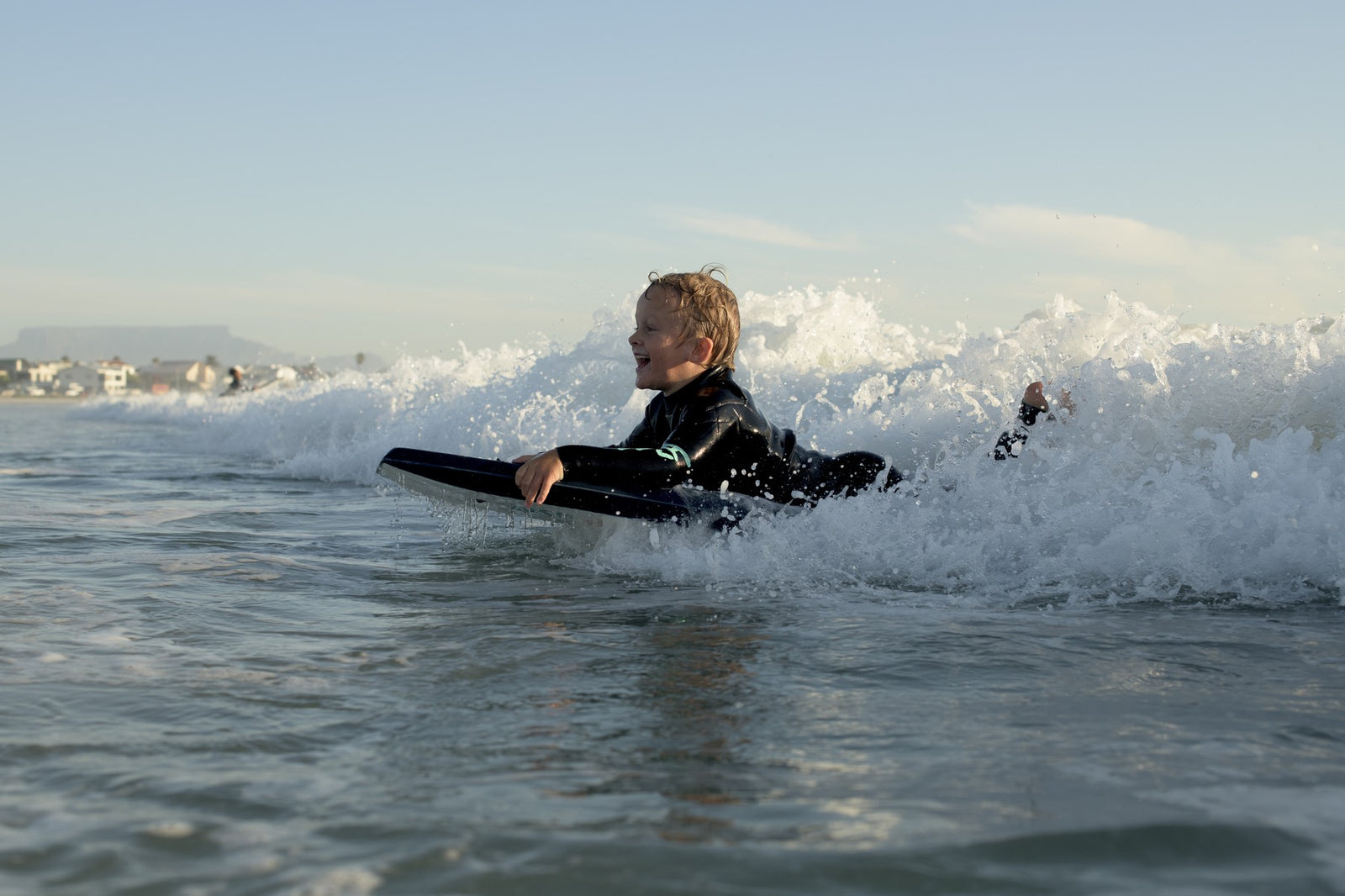A young boy in a wetsuit laughing as he rides a bodyboard through whitewash, Cape Town's coastline behind him