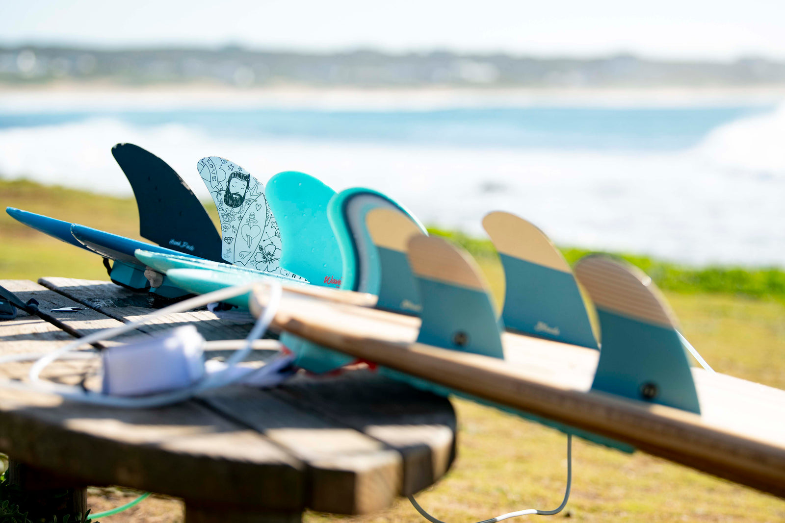 A row of surfboard fins, including a hand-drawn design, lined up on boards on a wooden table overlooking the sea