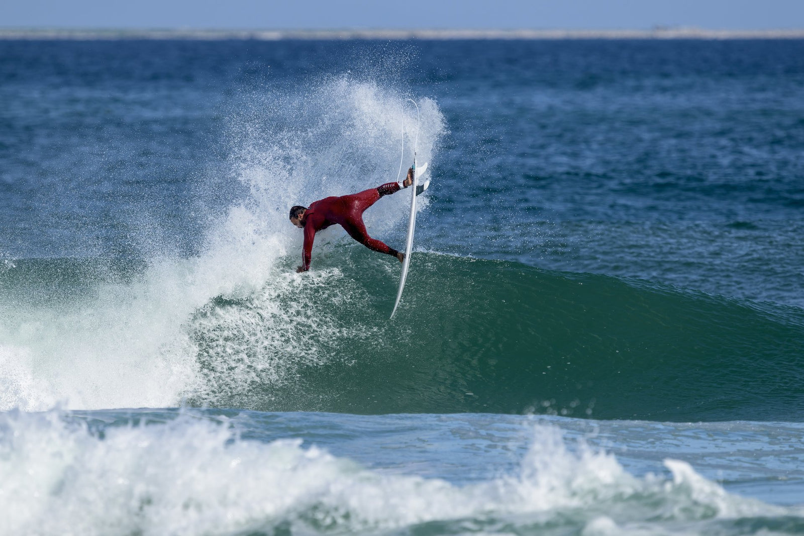 A surfer in a maroon wetsuit slashing off the lip of a green wave, board nearly vertical