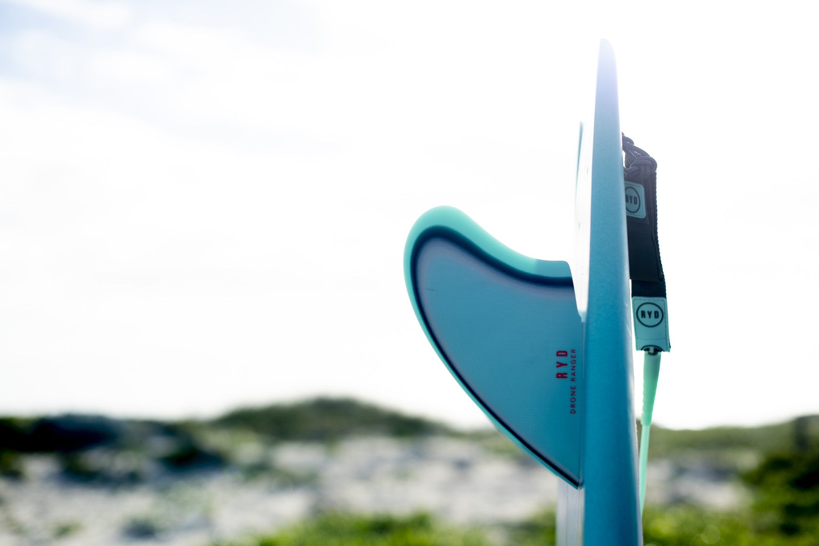 Close-up of a twin-fin surfboard's tail showing its teal fin, dunes blurred in the background