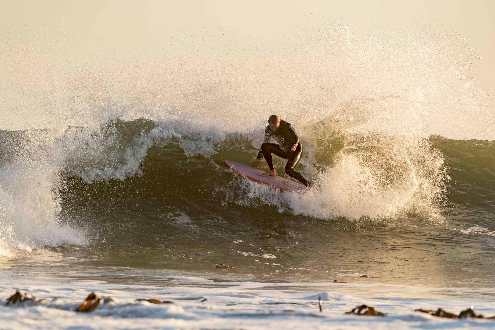 A surfer crouched low carving through a barrelling green-gold wave at sunset on a twin-fin board
