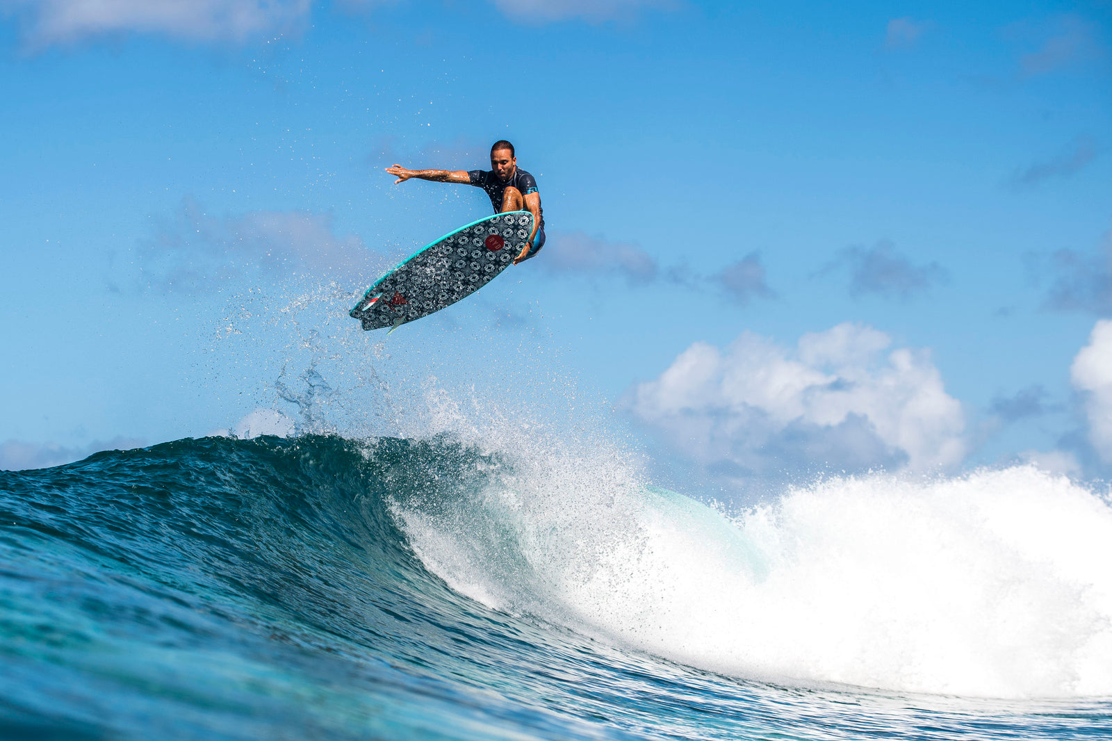 A surfer catching air above the lip of a wave on a patterned surfboard, silhouetted against a blue sky