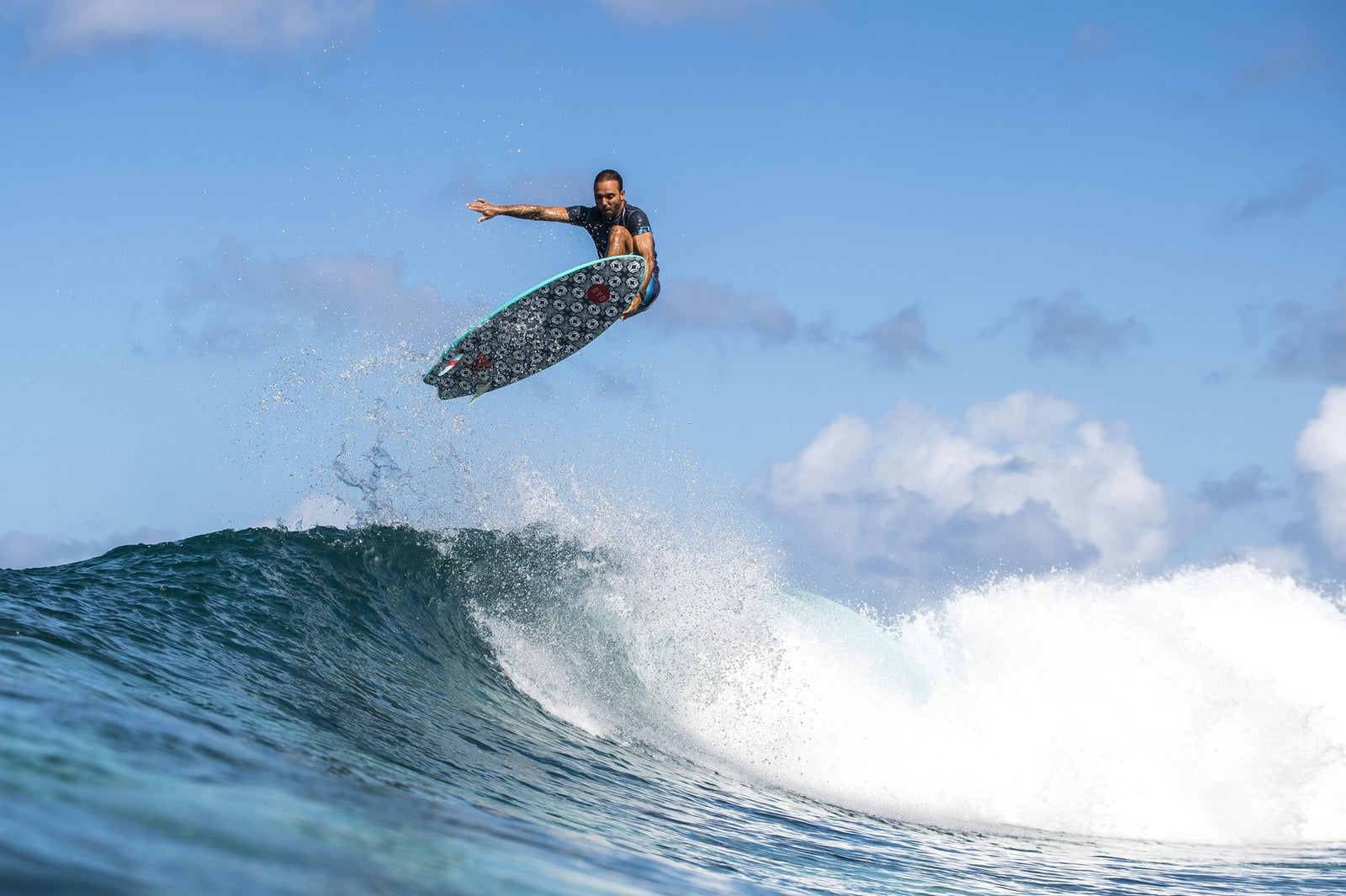 A surfer catching air above a breaking wave on a patterned softboard, framed against a bright blue sky