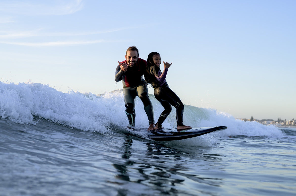 A father and daughter riding the same wave together on a black softboard, both giving the shaka hand sign