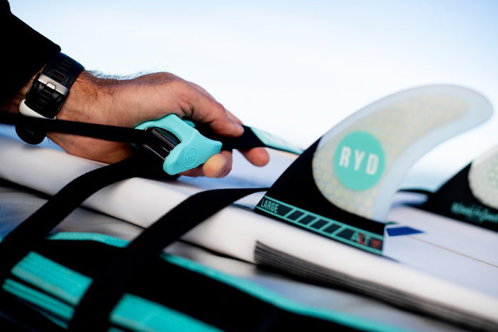 Hand tensioning a black RYD tie down strap with a teal cam buckle, securing a surfboard