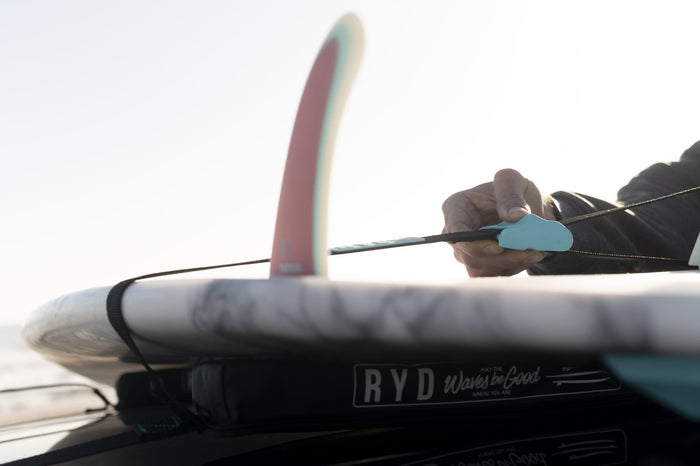 Hand tensioning a teal cam hook on a strap securing a marble-pattern surfboard to a soft roof rack