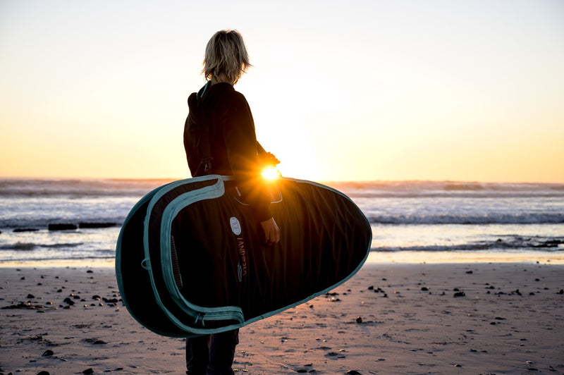 A surfer carrying an RYD Alt Range Everyday board cover at sunset