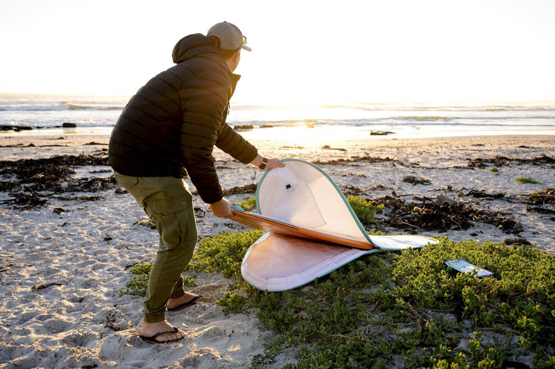 A surfer carrying an RYD Alt Range Simple board cover, looking out at the waves at sunset