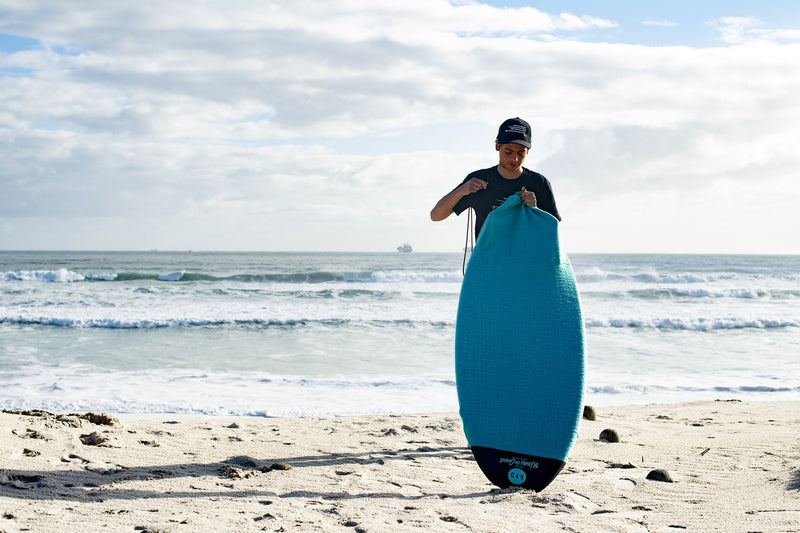 Pulling a teal RYD Alt Range Stretch board cover over a surfboard on the beach