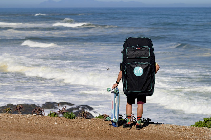 An RYD Bodyboard Range Everyday bag worn on the back, skateboard in hand, looking out at the waves