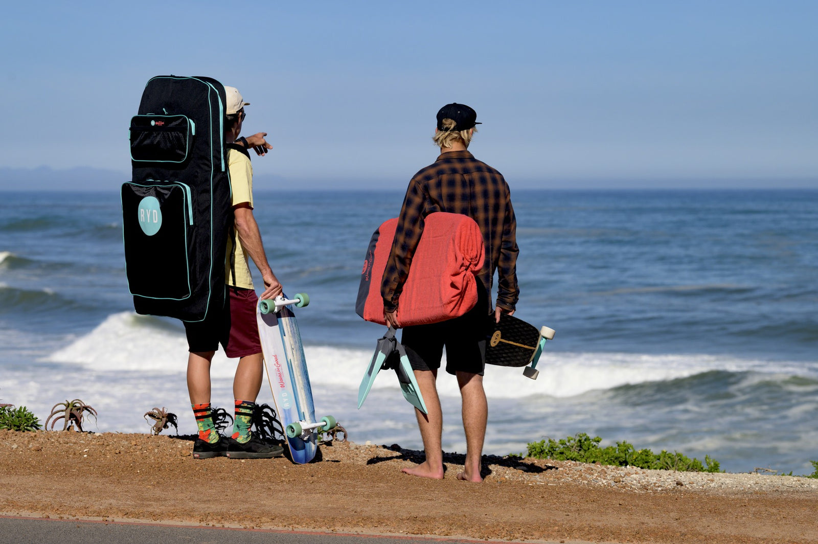 Two friends carrying an RYD board cover, fins, and skateboards, looking out at the waves before heading down to the beach