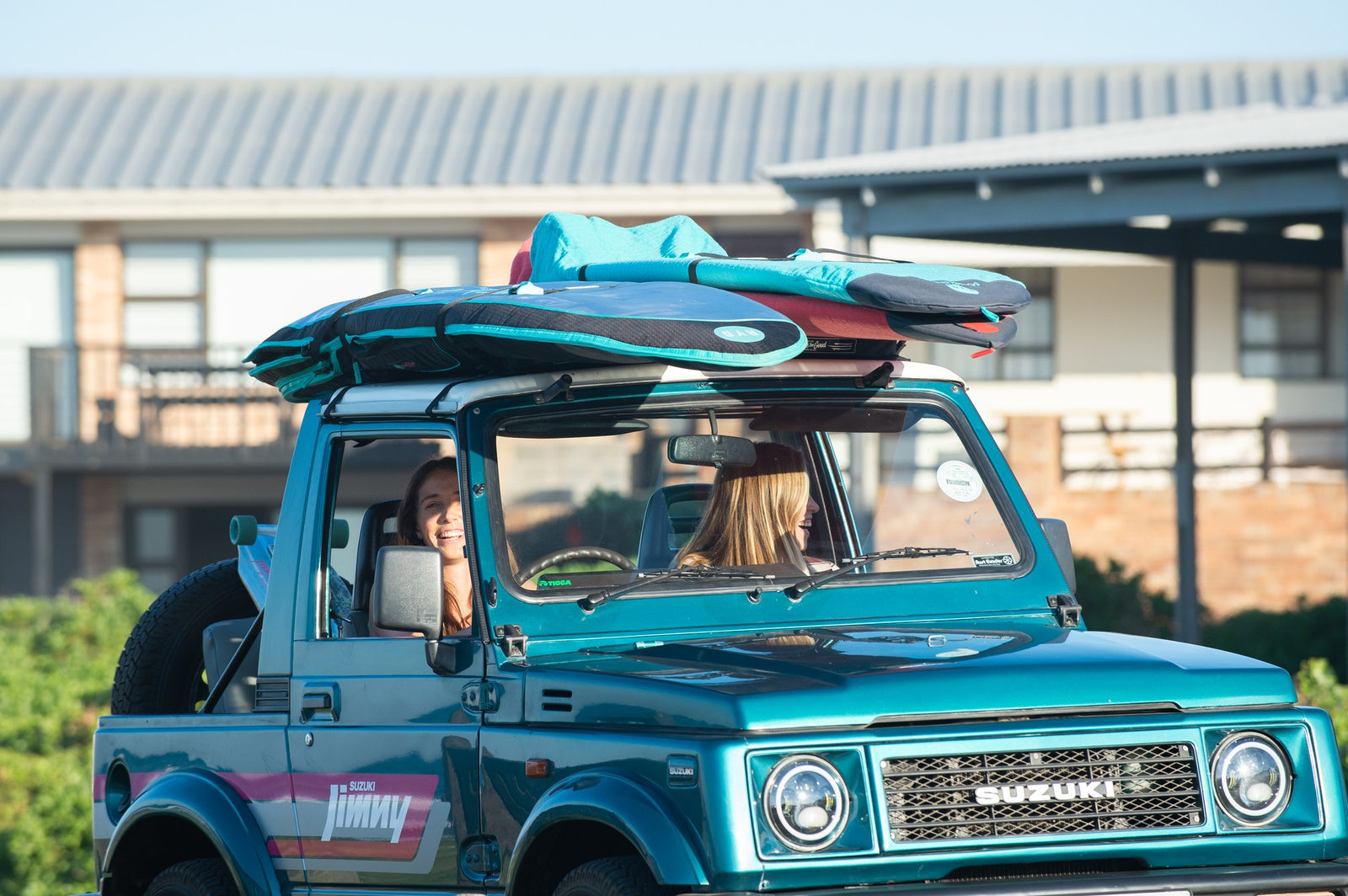 Two friends laughing inside a teal Suzuki Jimny with an RYD board cover and other board bags strapped to the roof rack, ready for a surf trip