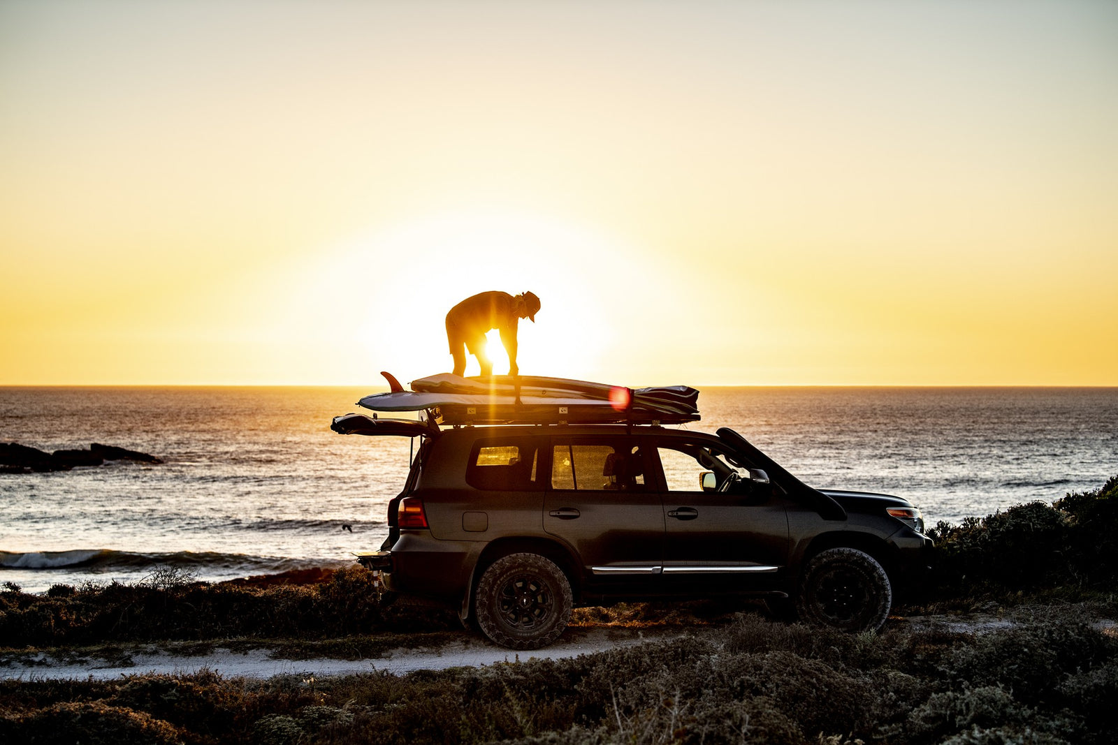 A surfer silhouetted against the sunset, strapping a board in its RYD cover onto the roof rack of a 4x4 parked above the ocean
