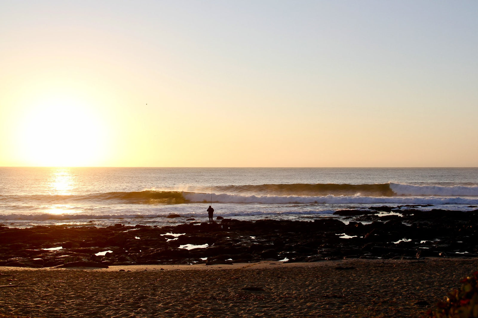 A lone fisherman silhouetted on rocks at sunset, casting a line into the sea as a wave breaks in the distance