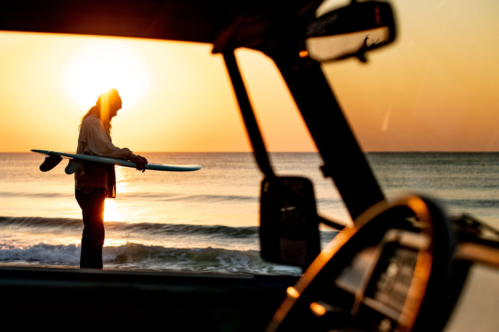 A surfer silhouetted at sunset, carrying a surfboard under one arm, framed through the open door and side mirrors of a vehicle with the ocean glowing gold behind him