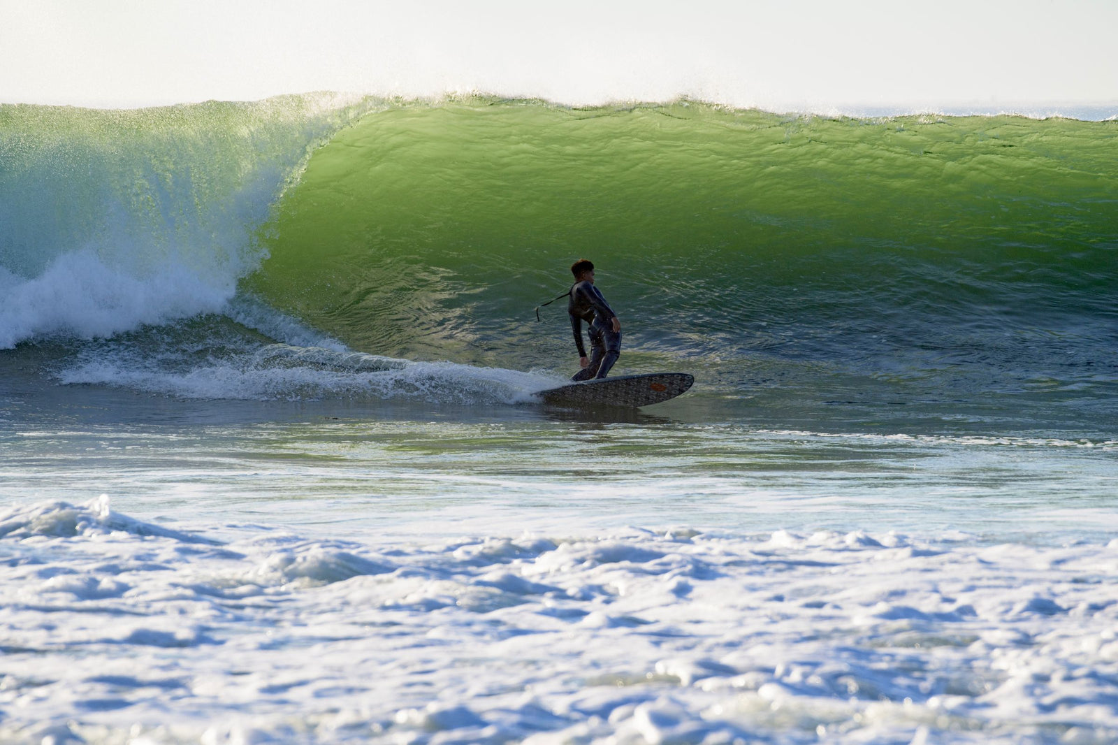 A silhouetted longboarder cross-stepping toward the nose of the board on a large sunlit wave