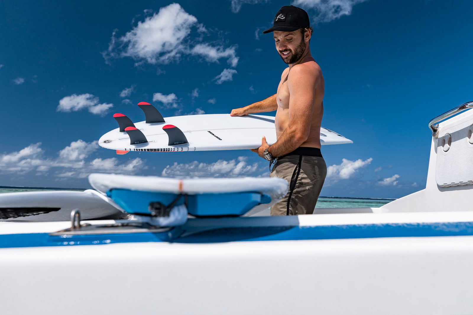A man carrying a white surfboard fitted with red-tipped quad fins across a boat deck
