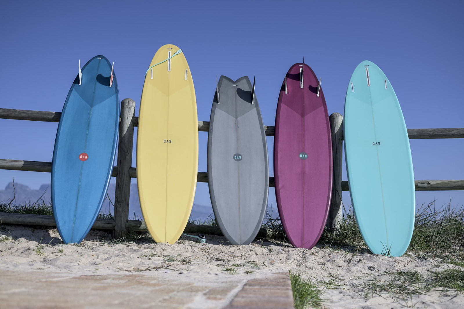 Five colourful surfboards leaning against a wooden fence in the sand, mountains in the distance