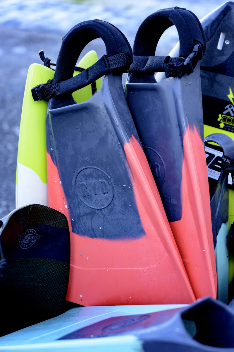 Two pairs of RYD swim fins in navy and coral, close up against a bodyboard and other beach gear