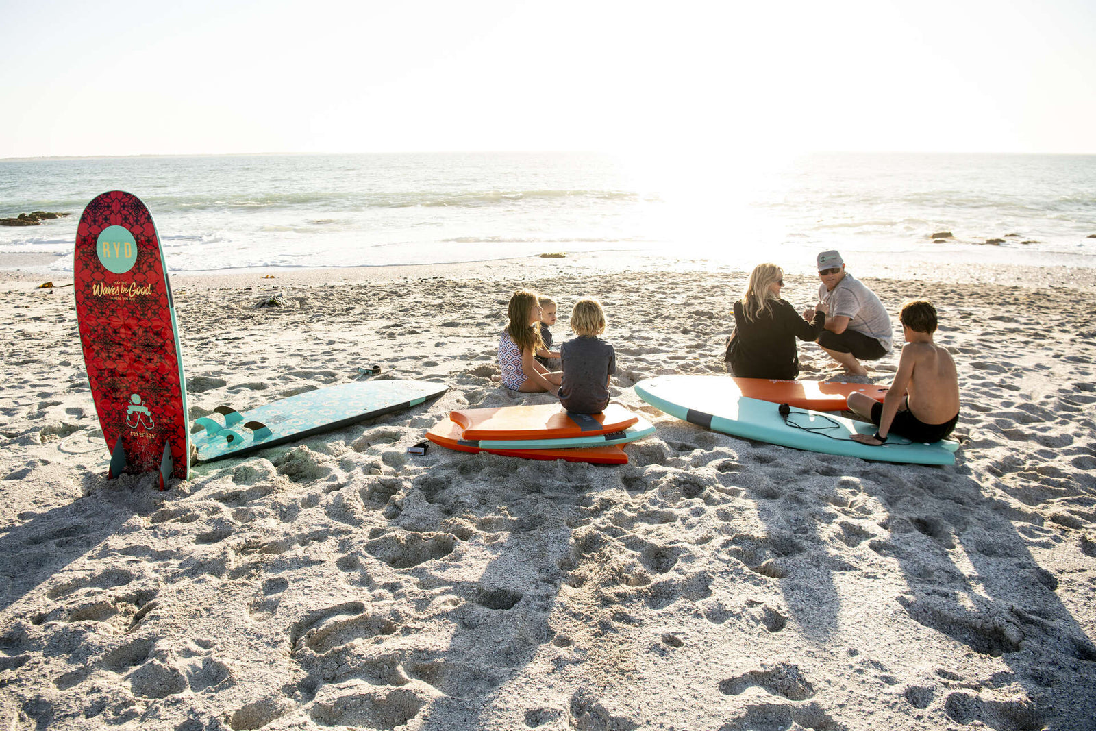A family sitting together on a sunny beach beside a row of surfboards and bodyboards planted upright in the sand