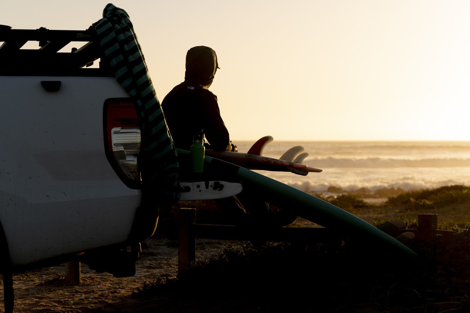 A surfer in a hooded jacket sitting on the open tailgate of a bakkie at sunset, resting a surfboard across his lap showing its fin setup