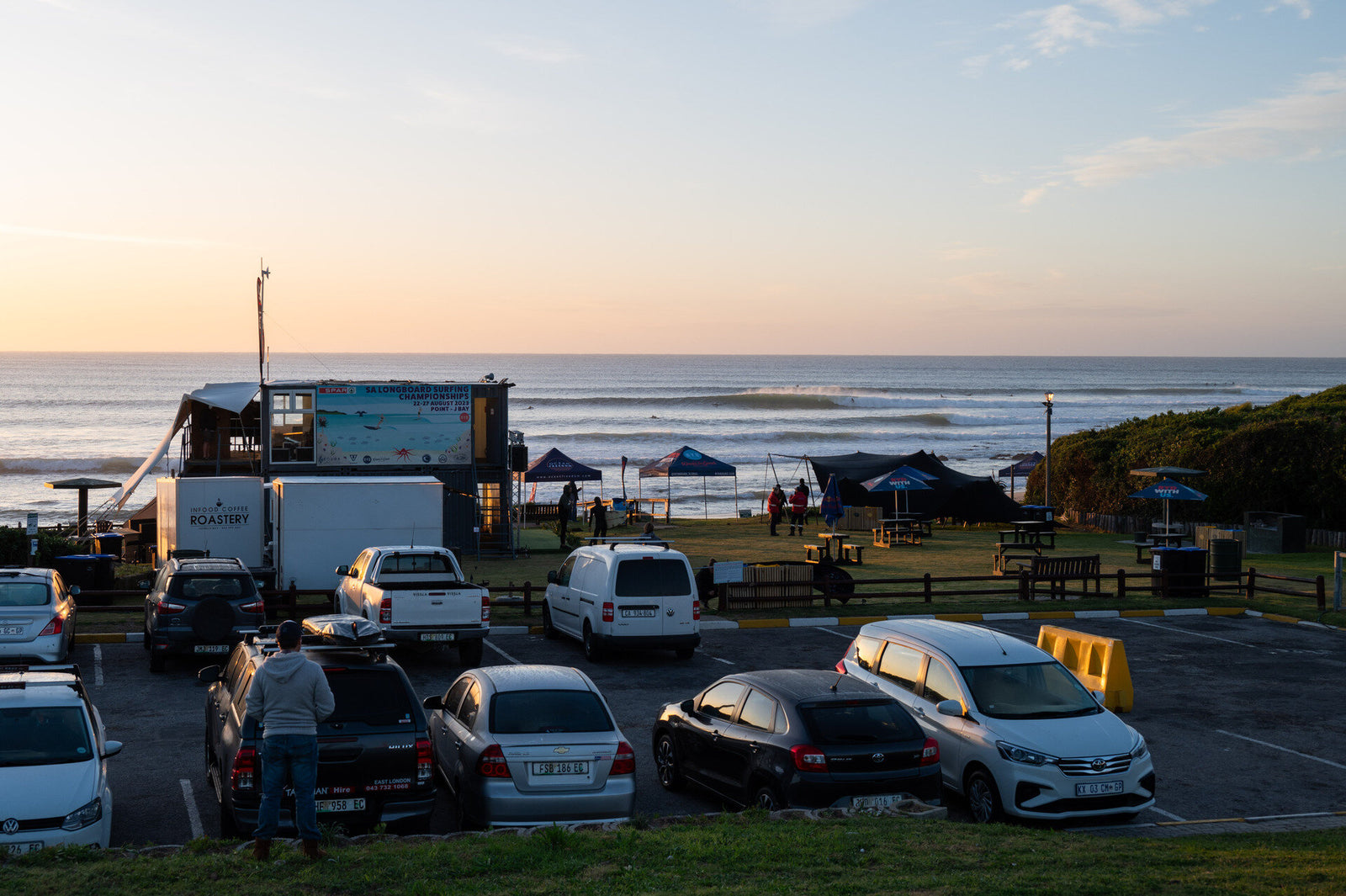 A car park overlooking a surf contest at sunset, with an event marquee for the SA Longboard Surfing Championships and surfers riding a wave in the background