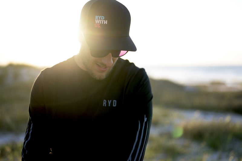 A man wearing a black RYD 'Ryd With Us' cap and black long-sleeve RYD top, backlit by golden hour sun on a coastal dune