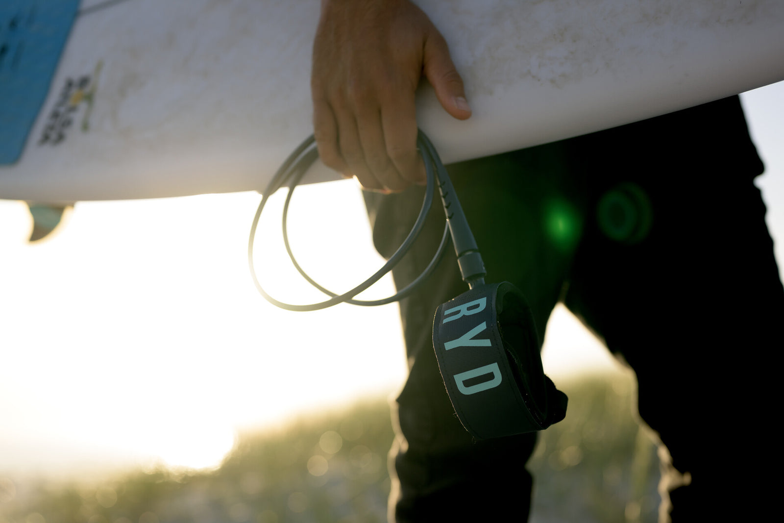 Close-up of a hand holding a coiled RYD leash by its ankle cuff, a white surfboard resting on the sand behind it