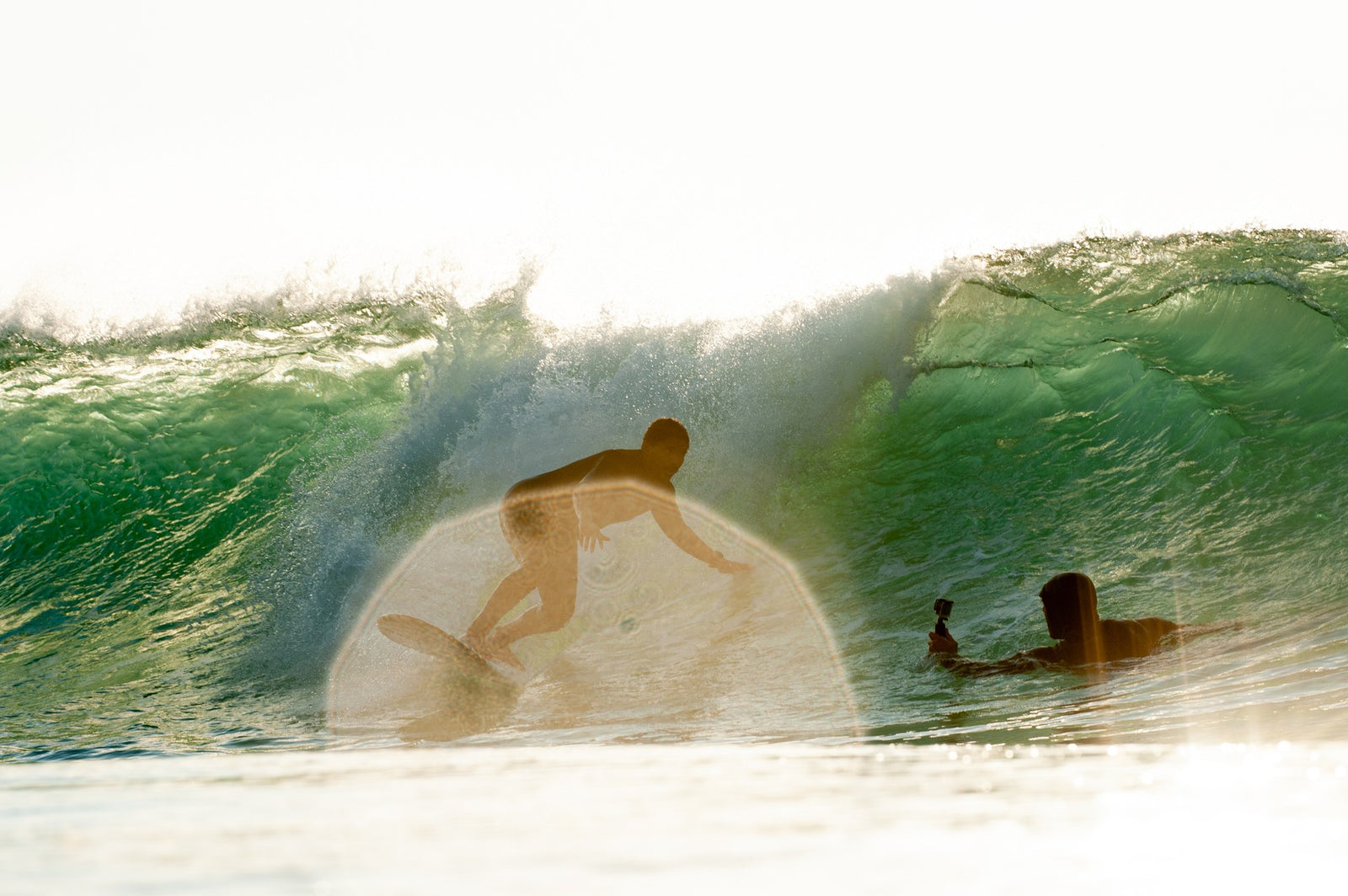 A surfer riding inside the barrel of a large green wave, with another swimmer holding a camera watching from the shoulder of the wave