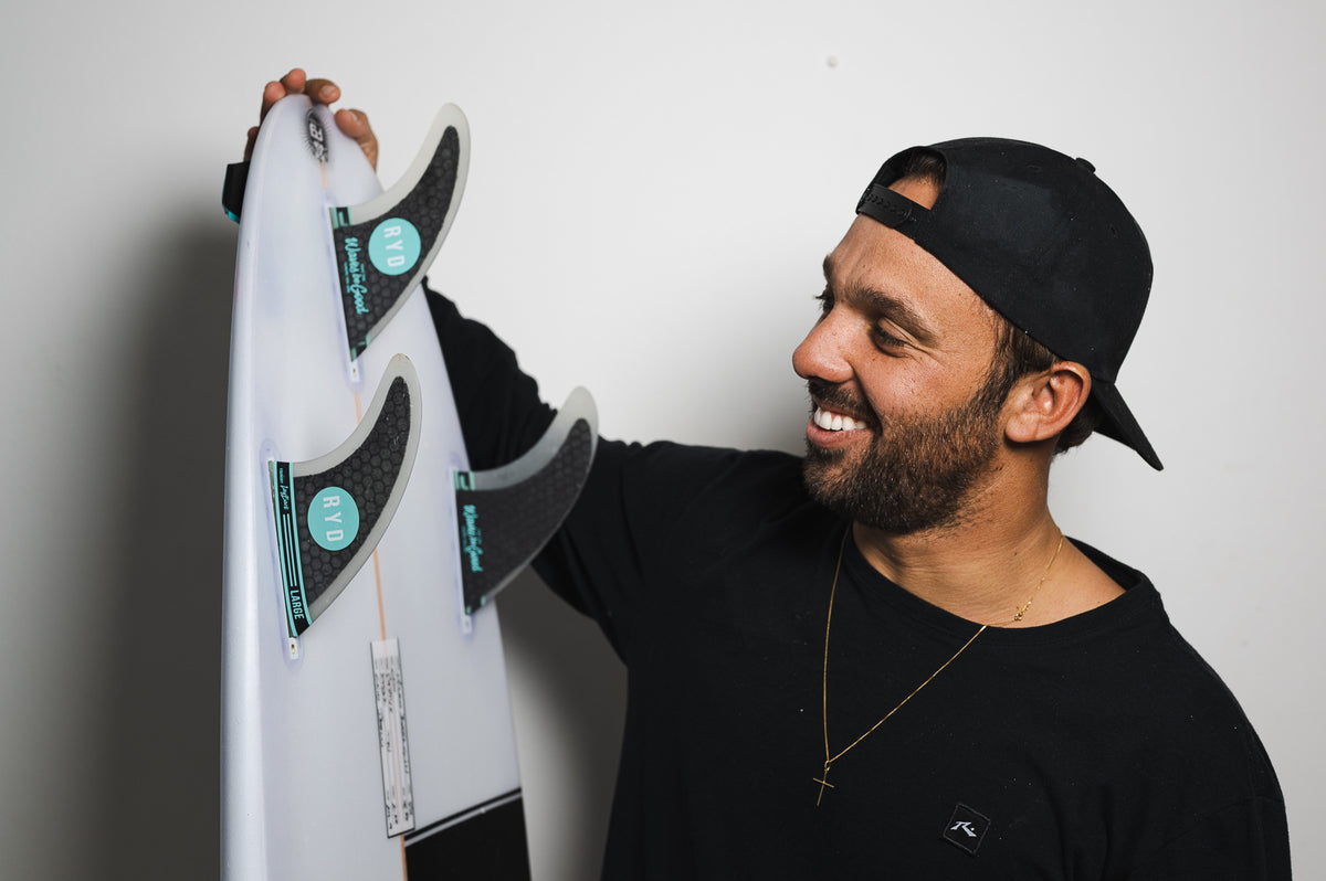 A smiling surfer holding up a white surfboard fitted with three RYD thruster fins, against a plain studio backdrop