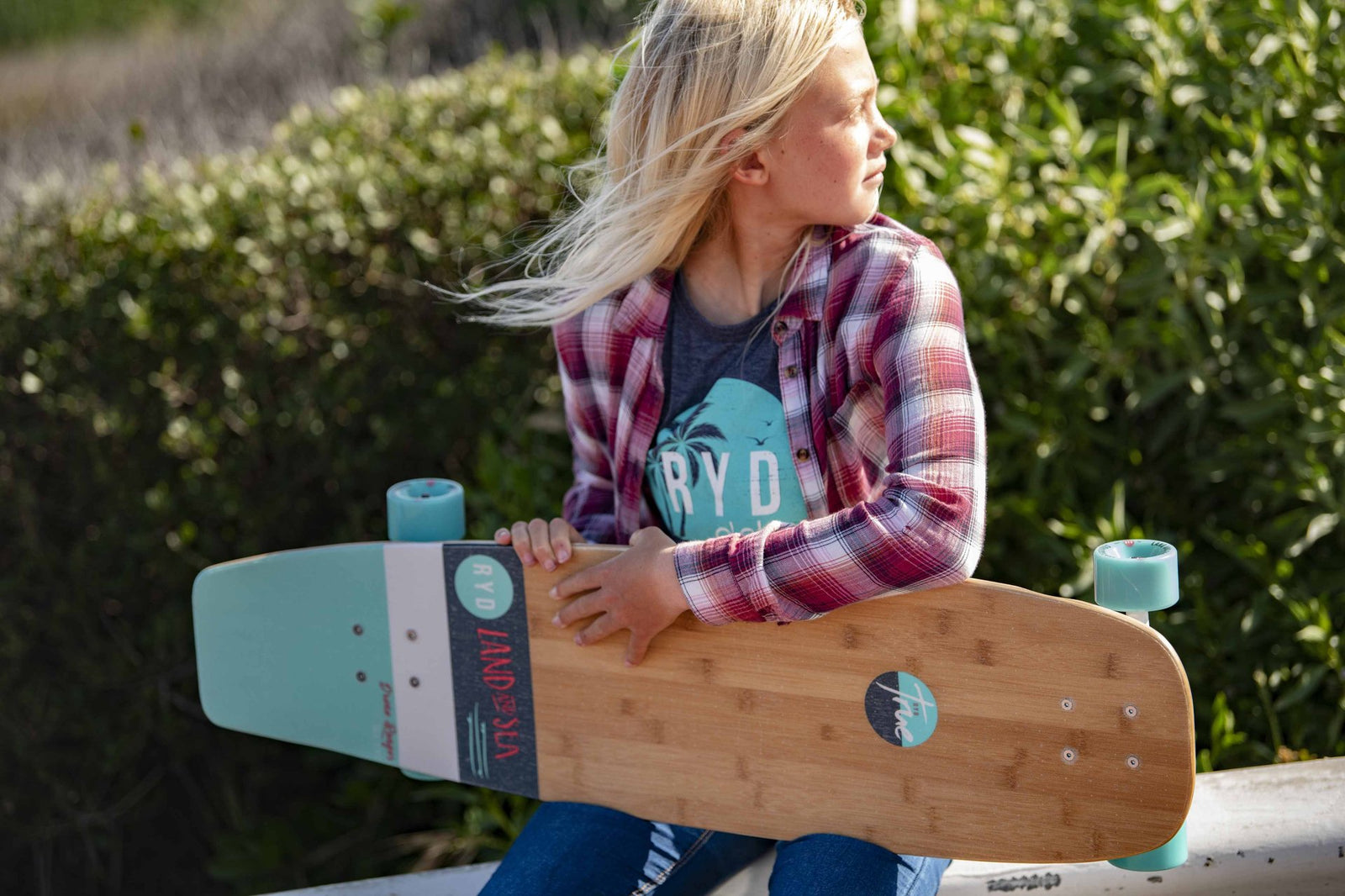A young girl with windswept blonde hair holding a wooden RYD Land and Sea cruiser skateboard with turquoise wheels, greenery behind her