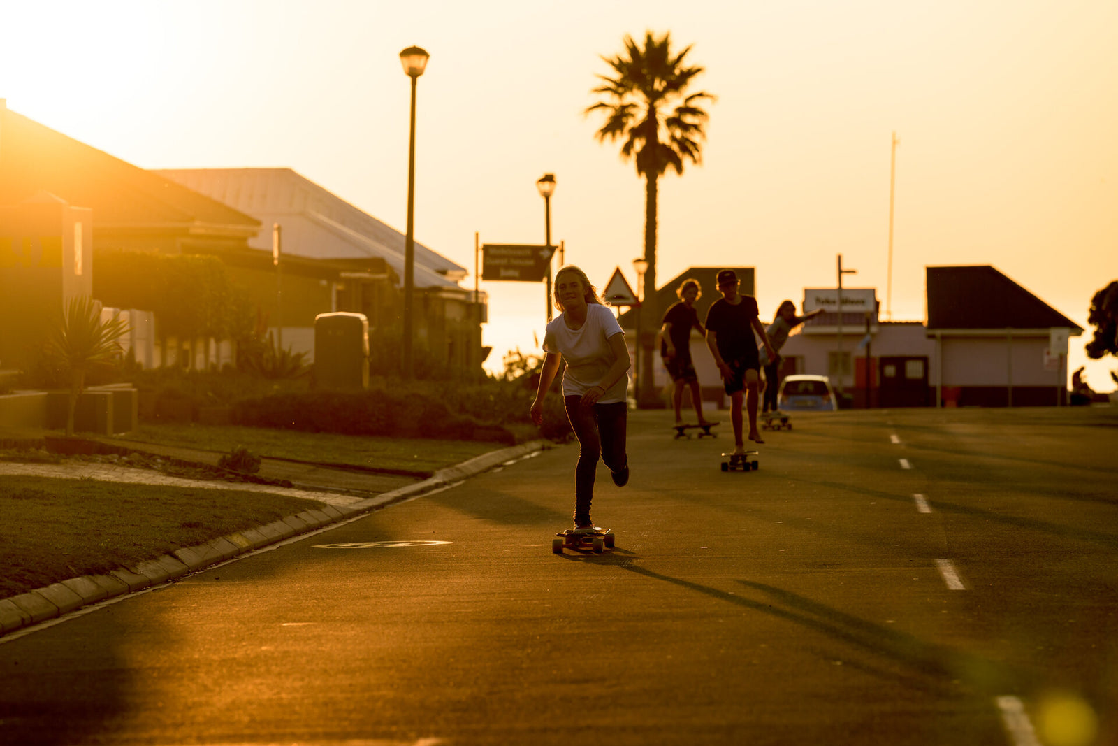 A group of friends cruising down a coastal street at sunset on RYD Retro Cruiser skateboards, palm tree and small-town buildings silhouetted against the golden sky