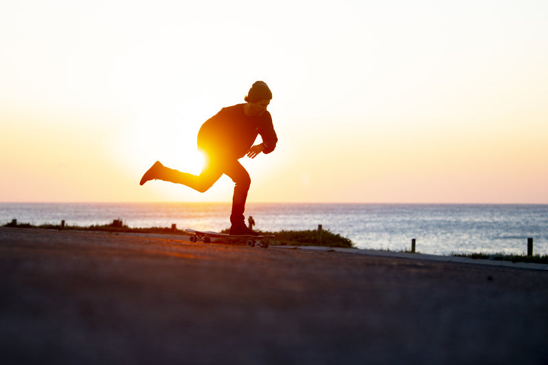 A skater silhouetted against a golden sunset, pushing along a coastal path with the ocean behind